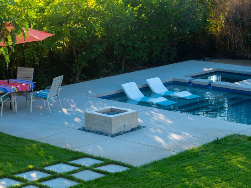 Contemporary backyard swimming pool with tanning ledge and white in-water lounge chairs surrounded by concrete decking and lawn.