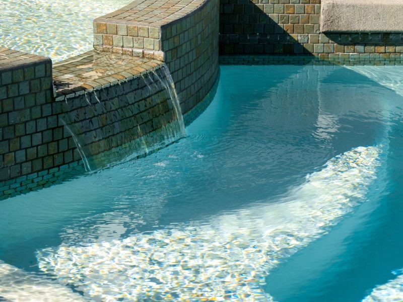 Close-up of tiled spa overflow waterfall flowing into clear blue swimming pool water.