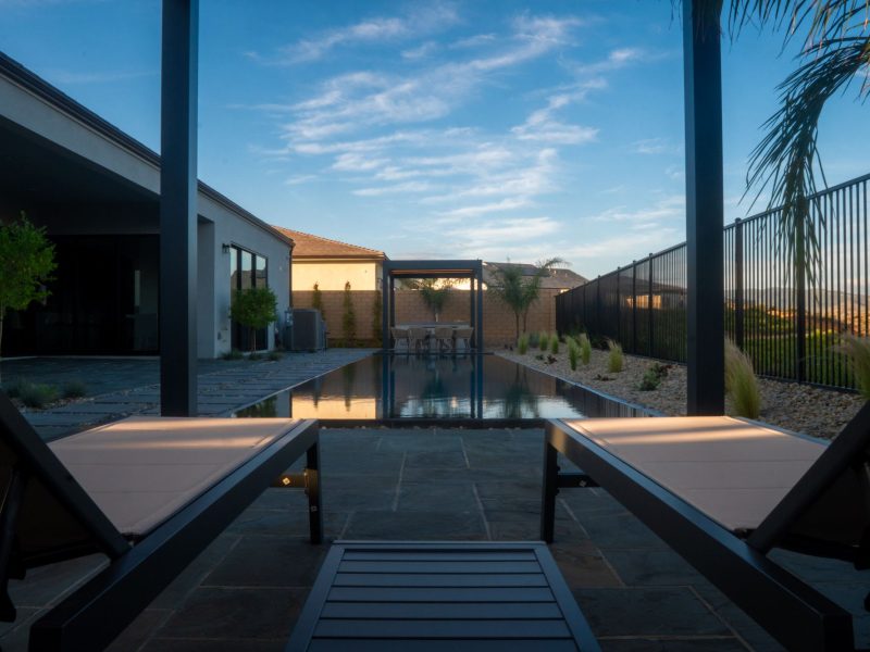 Two minimalist lounge chairs positioned under pergola looking toward long zero-edge reflecting pool and distant hillside landscape.