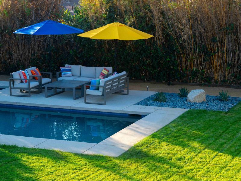 Modern poolside seating area with cushioned outdoor furniture, blue and yellow umbrellas, and concrete pool deck.