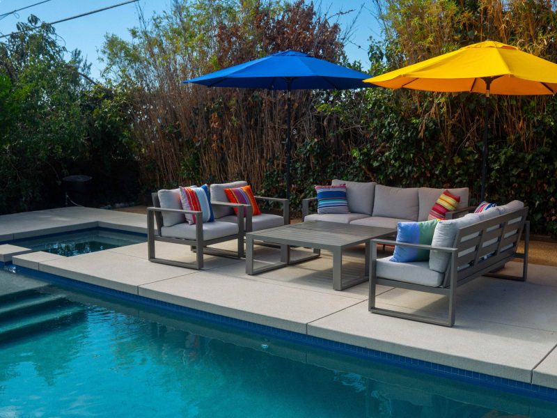 Cushioned outdoor sofa and chairs beside a residential swimming pool with umbrellas and concrete decking.