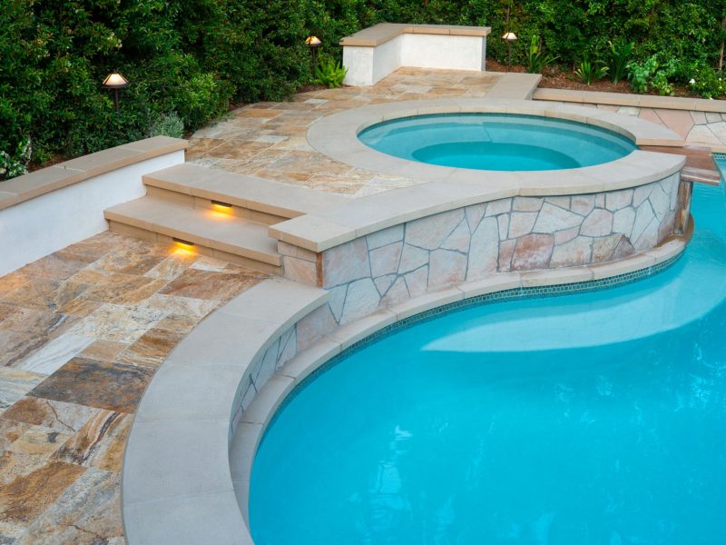 Circular raised spa with stone veneer surround, integrated steps, and adjacent swimming pool bordered by natural stone decking and landscape lighting.