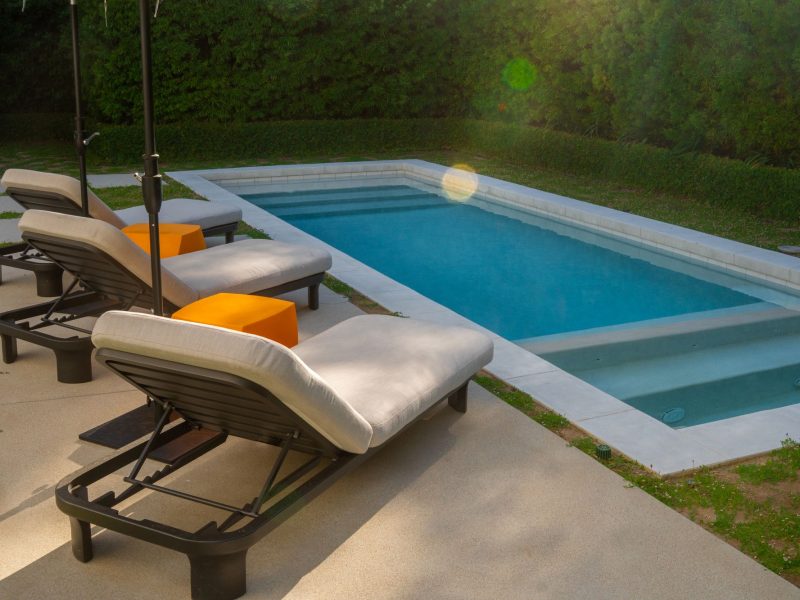 Minimalist rectangular pool bordered by concrete decking and cushioned lounge chairs in a modern backyard.