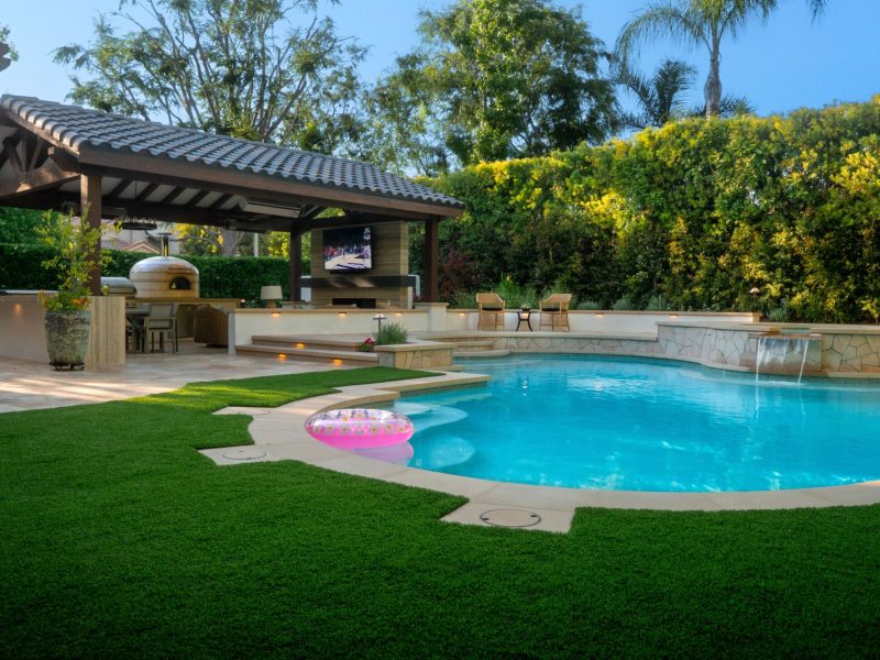 Curved swimming pool with spa, water features, green lawn, and covered entertainment area designed for relaxing and hosting gatherings outdoors.