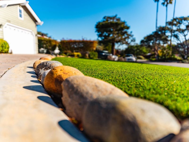 Close-up detail of artificial turf installation with decorative river rock border in a San Luis Obispo front yard.