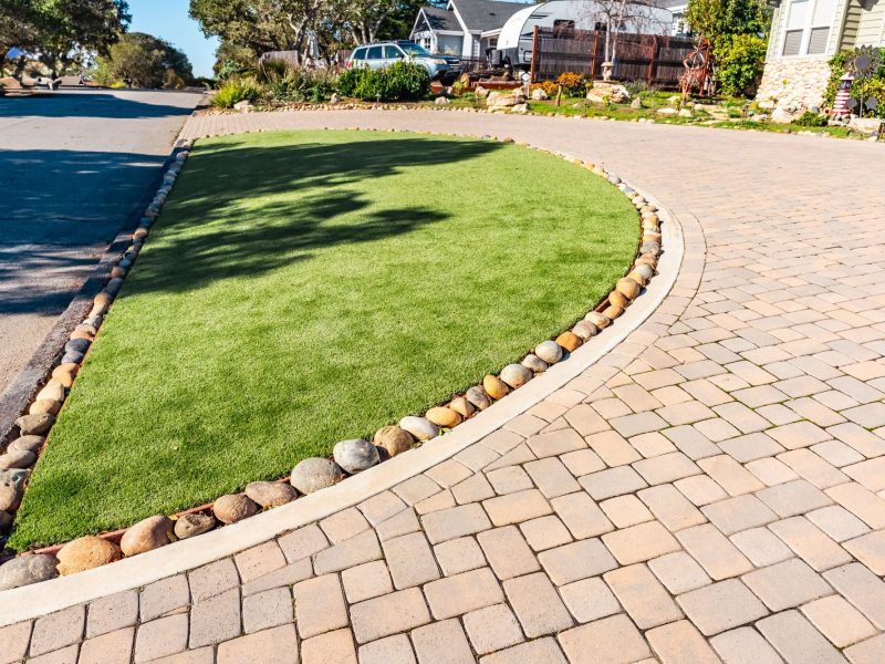 Curved artificial turf lawn in San Luis Obispo bordered by smooth river rocks and adjacent paver driveway.