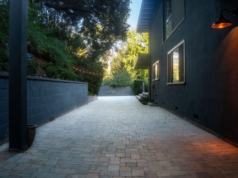 Long paver walkway along a contemporary dark-painted home with retaining wall, exterior lighting, and surrounding trees forming a clean modern landscape pathway.