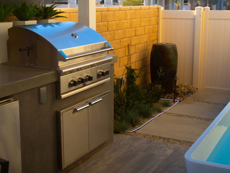 Compact outdoor living design featuring a built-in stainless steel grill, concrete countertop, and modern plunge pool beside landscaped planting.
