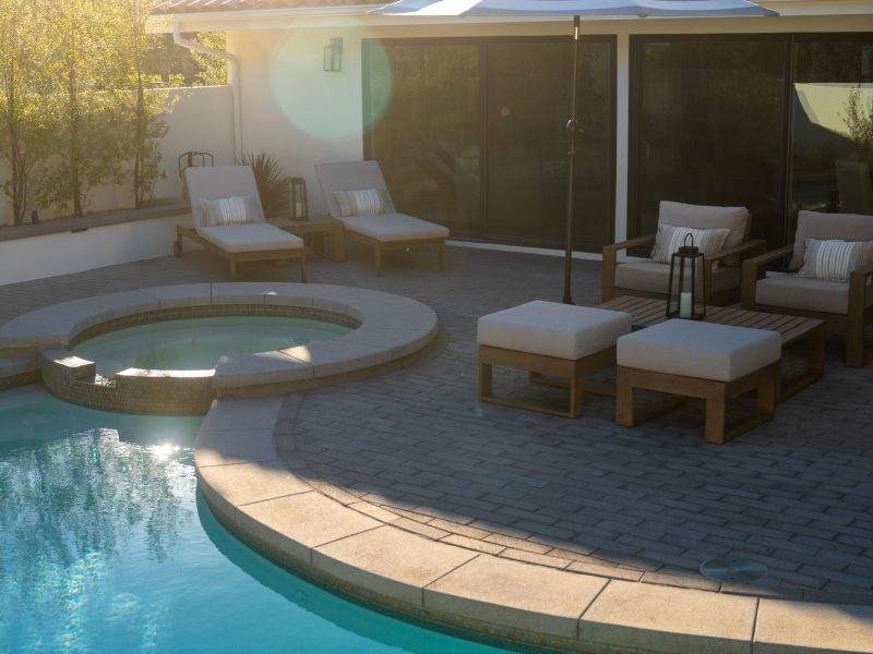 Raised spa connected to swimming pool with cushioned lounge chairs, umbrella, and paved patio in a contemporary backyard.