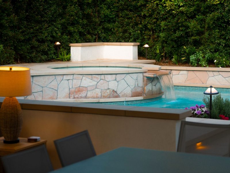 Raised spa with cascading waterfall spilling into illuminated swimming pool, surrounded by lush greenery and warm landscape lighting.