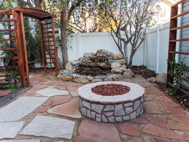 Custom backyard flagstone patio featuring a circular stone fire pit and natural rock waterfall water feature.