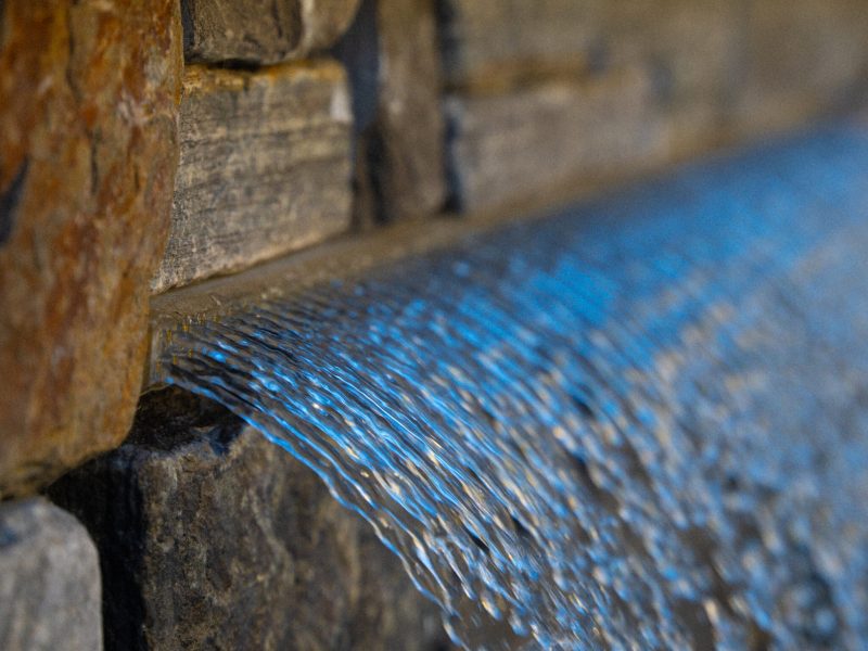 Water flowing from a stone wall scupper into a residential swimming pool, creating a decorative backyard water feature.