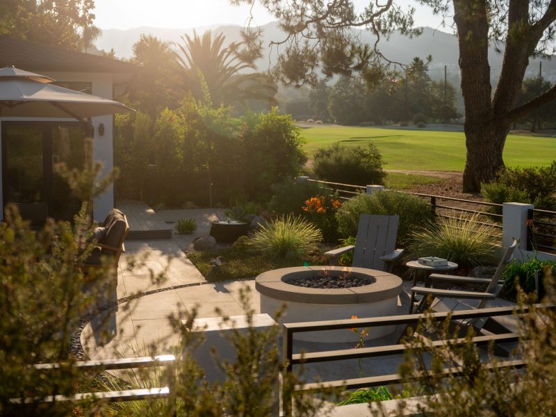 Warm sunset lighting over a landscaped backyard patio with circular fire pit and expansive golf course scenery.