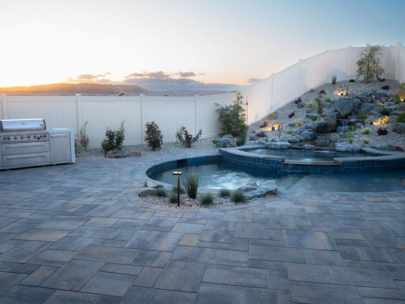 Backyard pool and raised waterfall feature beside a built-in stainless outdoor kitchen, photographed at sunset with warm sky tones.