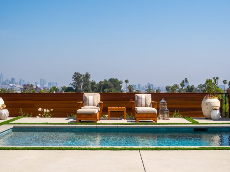 Balanced arrangement of outdoor lounge chairs facing a modern swimming pool with panoramic city skyline beyond a wood privacy fence.