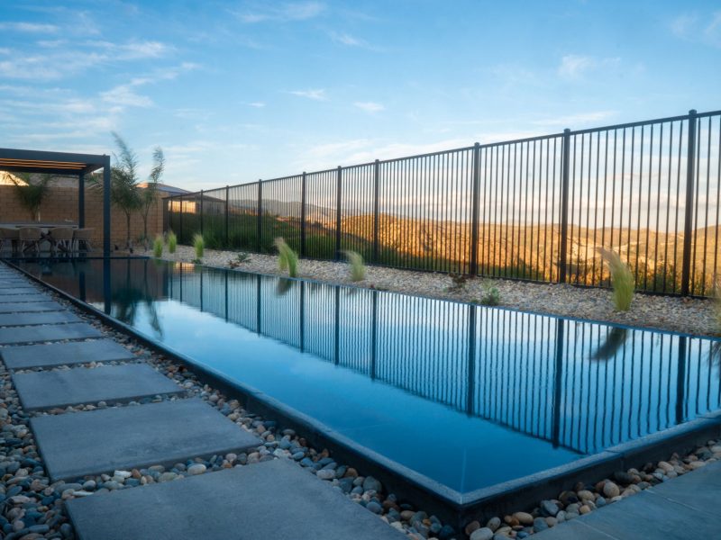 Long rectangular zero-edge pool bordered by stepping stone walkway, river rock, and black metal fencing overlooking scenic hillside views.